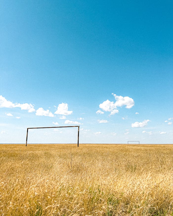 A wide view of a rural soccer field with goal posts in a grassy landscape under a blue sky.